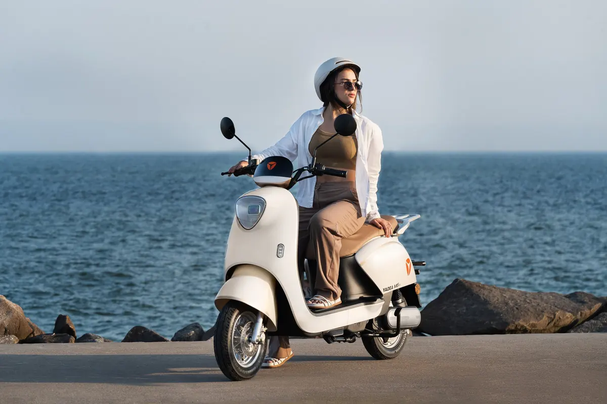 Mulher de capacete branco pilotando uma scooter elétrica Yadea M6 num calçadão à beira-mar